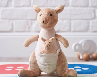 May include: A plush kangaroo toy in shades of tan and beige. The larger kangaroo has a pouch with a smaller kangaroo inside. The toy is soft and cuddly, with a friendly expression. The background includes a white brick wall and a colorful rug.