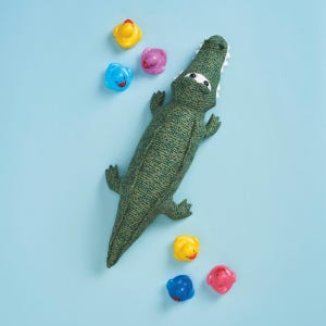May include: A green fabric alligator toy with white teeth and eyes, surrounded by colorful plastic bath toys. The alligator is textured and detailed, lying on a light blue surface. The bath toys include yellow, blue, and pink ducks and other animal shapes.