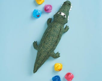 May include: A green fabric alligator toy with white teeth and eyes, surrounded by colorful plastic bath toys. The alligator is textured and detailed, lying on a light blue surface. The bath toys include yellow, blue, and pink ducks and other animal shapes.