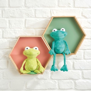 Pode incluir: Dois brinquedos de pelúcia de sapo em um ambiente de berçário. Um é verde limão, o outro é azul-turquesa. Eles estão em prateleiras hexagonais de madeira com fundos rosa e verde, montadas em uma parede de tijolos brancos. Os sapos têm olhos grandes e estão sorrindo.