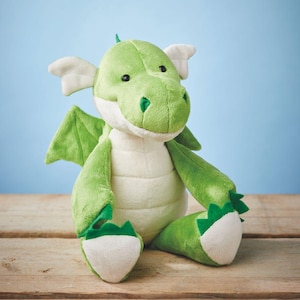 Könnte beinhalten: Ein grünes Plüschdrachen-Spielzeug mit weißen Akzenten an Flügeln, Bauch und Füßen. Der Drache hat schwarze Augen und ein kleines grünes Horn. Das Spielzeug sitzt auf einer Holzoberfläche vor blauem Hintergrund. Ein weiches Spielzeug für Kinder.