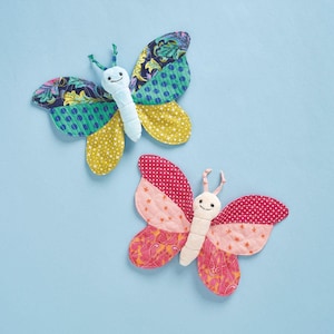 Può includere: Due peluche a forma di farfalla fatti a mano. Uno ha ali blu, verdi e gialle, l'altro rosa e arancioni. Entrambi hanno facce sorridenti e antenne, su uno sfondo azzurro.