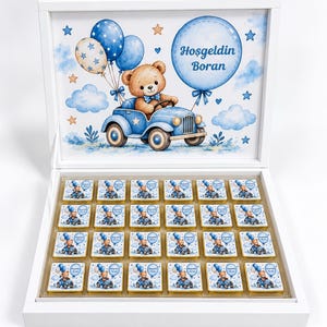 Könnte beinhalten: Eine weiße rechteckige Schachtel mit vierundzwanzig quadratischen Pralinen, die jeweils in Papier mit einem Teddybären in einem blauen Auto und blauen Luftballons eingewickelt sind. Der Innendeckel zeigt das Motiv mit dem Text Hoşgeldin Boran.