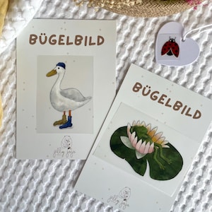 Könnte beinhalten: Zwei Bügelbilder auf weißen Karten. Das erste Design zeigt eine weiße Ente mit einem blauen Hut und blauen Stiefeln. Das zweite Design zeigt einen grünen Frosch, der auf einem rosa Seerosenblatt schläft. Beide Karten haben den Text "Bügelbild" und das Logo "jelly jojo" aufgedruckt.