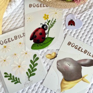 Könnte beinhalten: Drei Bügelbilder mit weißem Hintergrund. Ein Bügelbild zeigt einen Marienkäfer auf einem grünen Blatt mit weißen Blüten. Ein weiteres Bügelbild zeigt ein graues Kaninchen mit einem gelben Schal. Das letzte Bügelbild zeigt weiße Blüten und grüne Blätter.