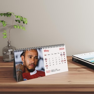 May include: A desk calendar for May 2026 featuring a portrait of a man in a red shirt. The calendar has a spiral binding and displays the days of the month with the US flag next to the dates. The text "May" and "2026" are printed in red.