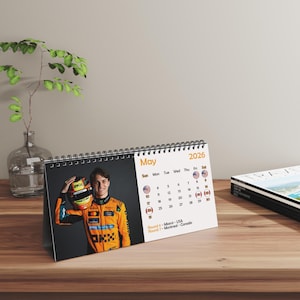 Puede incluir: Un calendario de escritorio para mayo de 2026 con un piloto de Fórmula 1 con un traje de carreras naranja. El calendario muestra los días del mes con las ubicaciones de las carreras. El calendario está sobre una superficie de madera.