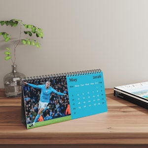 Puede incluir: Un calendario de escritorio para mayo de 2026 con un jugador de fútbol con una camiseta azul claro. El calendario tiene un fondo azul con los días de la semana y las fechas. El calendario está sobre una superficie de madera.