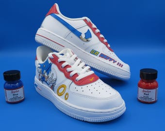 May include: White sneakers with red and blue accents, featuring a cartoon character design. The shoes have white laces and soles, with the text "GO HAPPY III" on the side. Two small bottles of acrylic leather paint, blue and red, are also in the image.