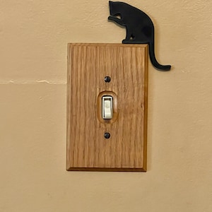 May include: A light switch cover made of wood with a black metal cat silhouette on top. The cat is in a sitting position with its tail curled around the cover.
