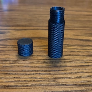 May include: A black, textured, cylindrical object with a threaded top and a separate cap. The cap and body have a diamond pattern. The objects are likely components of a larger item.