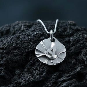 Peut inclure: Un collier pendentif argenté avec un pendentif circulaire au design texturé et superposé, orné d'une petite silhouette d'oiseau. Le pendentif est suspendu à une fine chaîne en argent. Le collier est présenté sur une surface sombre et texturée.