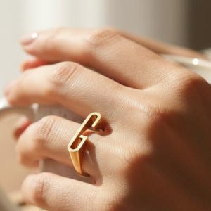 Könnte beinhalten: Ein goldfarbener Ring mit rechteckigem Design wird an einem Finger getragen. Der Ring ist der Mittelpunkt, vor dem Hintergrund von Händen, die eine weiße Tasse halten. Die Szene ist in natürliches Licht getaucht, was eine warme, einladende Atmosphäre suggeriert.