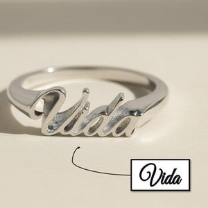 Könnte beinhalten: Ein silberner Ring mit dem Wort "Vida" in Schreibschrift. Der Ring liegt auf einer hellbeigen Oberfläche, mit einem kleinen Textfeld mit "Vida" in Schreibschrift. Der Ring hat ein schlichtes Banddesign.