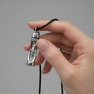 Könnte beinhalten: Eine silberfarbene Anhängerkette mit schwarzem Band. Der Anhänger ist ein kleines, rechteckiges Fläschchen mit einer detaillierten Figur im Inneren. Die Kette wird vor einem schlichten grauen Hintergrund gehalten.