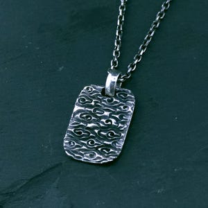 Può includere: Una collana con pendente color argento. Il pendente rettangolare presenta un motivo strutturato di linee a spirale. La catena è anch'essa argentata e ha un design a maglie. La collana è mostrata su uno sfondo scuro.