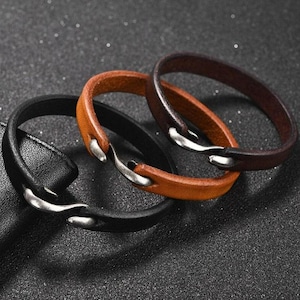 Könnte beinhalten: Drei Lederarmbänder in Schwarz, Braun und Beige. Jedes Armband hat einen silberfarbenen Verschluss. Die Armbänder sind auf einer dunklen, strukturierten Oberfläche angeordnet.