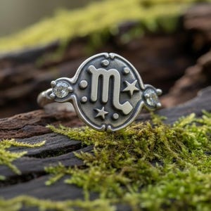 Op de afbeelding: Een zilveren ring met het sterrenbeeld Schorpioen. De ring heeft een cirkelvormig ontwerp met het symbool in het midden, geflankeerd door sterren en edelstenen. De ring is geplaatst tegen een achtergrond van mos en hout.