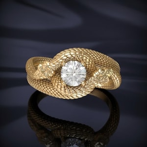 Op de afbeelding: Een gouden ring met een gestructureerd, schubachtig slangendesign dat om een enkele, ronde, heldere gefacetteerde edelsteen in het midden is gewikkeld. De band vertoont gedetailleerde slangenhuistexturen, waarbij twee slangenkoppen elkaar bij de steen ontmoeten.