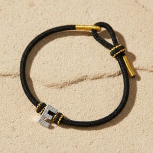 May include: Black rope bracelet with gold-colored accents and a silver letter "E" encrusted with small, clear stones. The bracelet is set against a sandy background, creating a contrast of textures and colors. A stylish piece of jewelry.