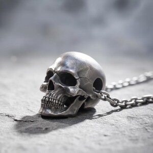 Puede incluir: Colgante de calavera plateada con cadena. La calavera presenta detalles como cuencas de los ojos, cavidad nasal y dientes. La cadena está hecha de eslabones pequeños e interconectados. El colgante descansa sobre una superficie gris texturizada.