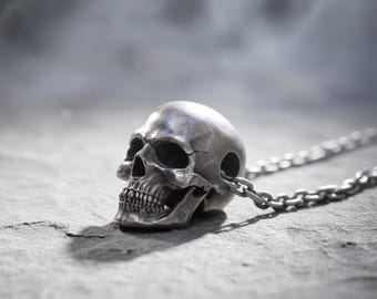 Könnte beinhalten: Silberner Totenkopf-Anhänger mit Kette. Der Schädel weist detaillierte Augenhöhlen, eine Nasenhöhle und Zähne auf. Die Kette besteht aus kleinen, ineinandergreifenden Gliedern. Der Anhänger liegt auf einer strukturierten, grauen Oberfläche.