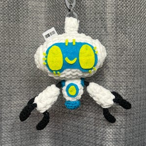 May include: A white crocheted robot keychain with a blue face, yellow eyes, and black arms and feet. The robot has a blue and yellow center and is attached to a silver keychain ring.