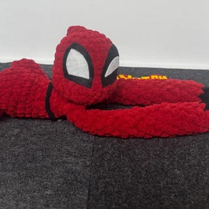 May include: A red plush toy in the shape of a superhero, with a black and white mask and black accents. The toy has long arms and legs, and a gray clasp at the end of one arm. The toy is lying on a gray surface.
