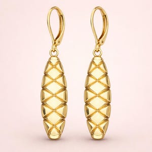 May include: A pair of gold drop earrings. Each earring has a long, geometric design with diamond-shaped patterns. The earrings have a lever-back closure. The earrings are displayed against a soft pink background.