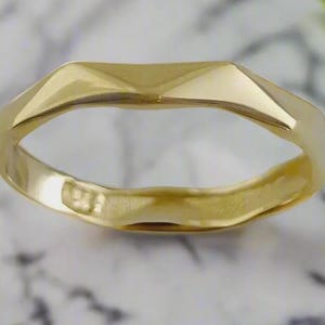 Puede incluir: Primer plano de un anillo de oro con un diseño geométrico. El anillo tiene una parte superior facetada y una banda lisa y pulida. El anillo está sobre un fondo borroso.