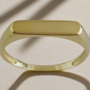 May include: A gold signet ring with a rectangular top. The ring is made of polished gold and has a smooth, rounded band. The ring is shown on a neutral background with shadows.