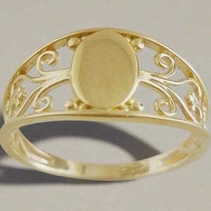 May include: A gold ring with an oval center and ornate filigree detailing. The ring has a polished finish and a delicate, openwork design. The band is smooth and the ring appears to be made of solid gold.