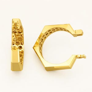 May include: Two gold-colored hoop earrings. One earring is shown from the side, revealing a honeycomb pattern on the inside. The other earring is shown from the front, with a geometric, hexagonal design. The earrings have a polished, reflective surface.