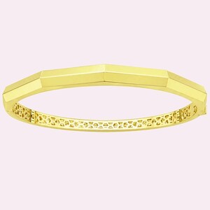 May include: A gold-colored bangle bracelet with a geometric design. The bracelet has a faceted top and a detailed, openwork pattern along the inner band. The overall design is modern and elegant.