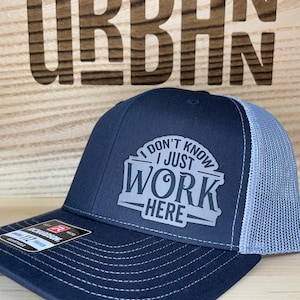 Puede incluir: Una gorra de camionero negra y gris con una malla gris en la parte posterior. La gorra tiene un diseño bordado en blanco que dice "No lo sé, solo trabajo aquí".