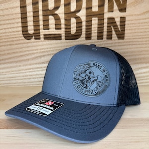 May include: A gray and black trucker hat with a curved brim. The hat features a circular patch with a skull graphic and the text "Hang in There, It Gets Worse." The hat is adjustable and has a mesh back.