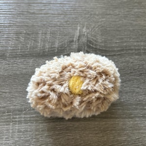 May include: A small, oval-shaped plush object covered in long, shaggy, beige-colored faux fur. The center of the plush features a small, circular, textured patch of bright yellow fabric, creating a distinct contrast against the surrounding beige fur.