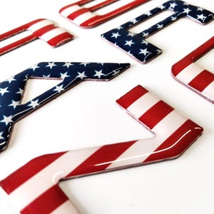 Puede incluir: Un conjunto de seis letras 3D, cada una con un acabado brillante en relieve. Las letras están pintadas con un diseño de bandera estadounidense roja, blanca y azul. Las letras están dispuestas en un orden aleatorio.