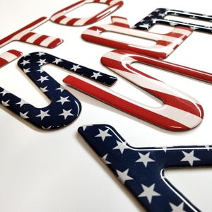May include: Close-up of several glossy, patriotic-themed letter stickers. The letters are in red, white, and blue, with some featuring the American flag design. The stickers are scattered on a white surface.