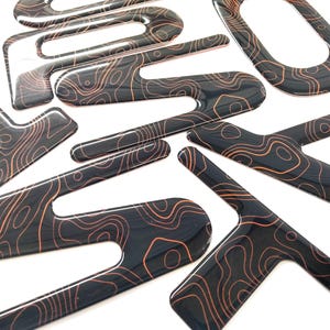 May include: A collection of glossy black decorative pieces with a topographic map design in shades of orange and brown. The shapes vary, including curved lines and geometric forms, all with a high-gloss finish.