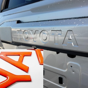 Puede incluir: Portón trasero plateado de coche con la palabra "TOYOTA" en relieve. Debajo, letras naranjas individuales deletrean "TOYOTA". Las letras tienen un acabado brillante. El fondo muestra un aparcamiento y gotas de agua.