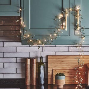 May include: A kitchen scene features a string of fairy lights with small green leaves, draped over a teal cabinet. The lights are strung along the cabinet's edge. Other items include a cutting board, a bottle, and a small potted plant.