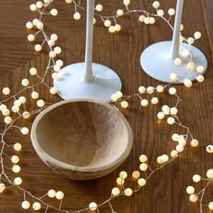 May include: A wooden bowl and two white stemmed glasses sit on a wooden surface, surrounded by a string of small, glowing, spherical lights. The lights are warm white and create a festive atmosphere. The bowl is light brown and has a natural, rustic look.