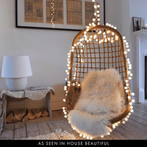 May include: A hanging rattan chair with a fluffy white cushion, adorned with fairy lights. A white lamp is on a small wooden table. The text "AS SEEN IN HOUSE BEAUTIFUL" is at the bottom of the image.
