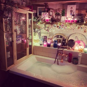 Puede incluir: Una ba&ntilde;era blanca llena de agua con espuma y un grifo de cobre. Junto a la ba&ntilde;era hay un armario de madera clara con puertas de cristal. La pared alicatada est&aacute; decorada con luces de hadas, velas, peque&ntilde;os cuadros y un letrero de Hello Kitty.