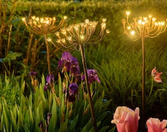 Könnte beinhalten: Gartendekoration mit drei beleuchteten Metallblumenskulpturen mit warmweißen Lichtern. Die Szene umfasst lila Iris, rosa Tulpen und grünes Gras und schafft so ein natürliches Ambiente. Die Lichter sind so gestaltet, dass sie Blütenköpfen ähneln.