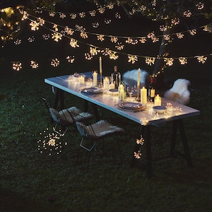 May include: A nighttime outdoor dining setting with a long wooden table set with plates, candles, and wine bottles. Snowflake-shaped string lights are draped overhead and around the table, creating a festive atmosphere. Two wire chairs with fur seat covers are visible.