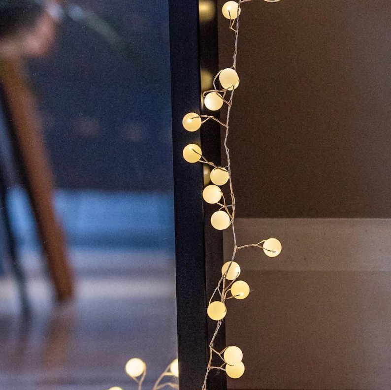 May include: String of warm white globe lights. The lights are strung on a thin, silver wire and are evenly spaced. The background is a dark brown and black.