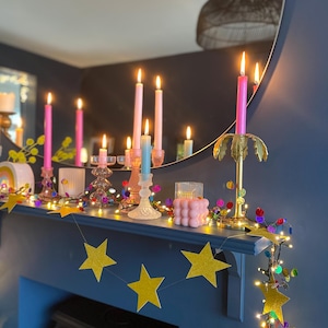 Puede incluir: Una colecci&oacute;n de velas c&oacute;nicas rosas y azules en portavelas de cristal y de palmera dorada, una vela de burbujas rosa y una guirnalda de estrellas doradas con purpurina y luces de colores sobre una repisa.