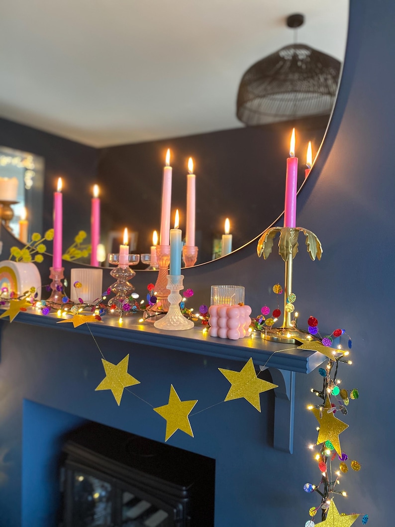Puede incluir: Una colecci&oacute;n de velas c&oacute;nicas rosas y azules en portavelas de cristal y de palmera dorada, una vela de burbujas rosa y una guirnalda de estrellas doradas con purpurina y luces de colores sobre una repisa.